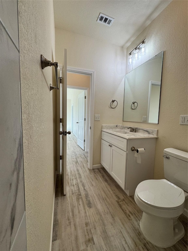 half bath with visible vents, toilet, wood finished floors, a textured wall, and vanity