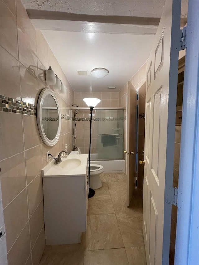 bathroom featuring visible vents, bath / shower combo with glass door, toilet, vanity, and tile walls