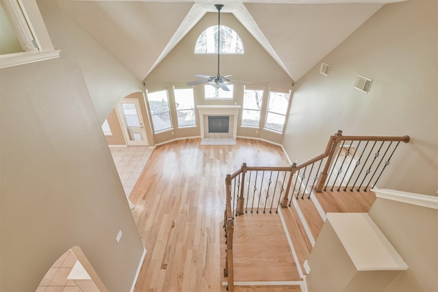 unfurnished living room with ceiling fan, stairway, wood finished floors, a glass covered fireplace, and high vaulted ceiling