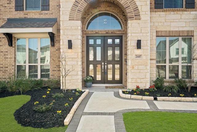 view of exterior entry featuring french doors, stone siding, brick siding, and a shingled roof