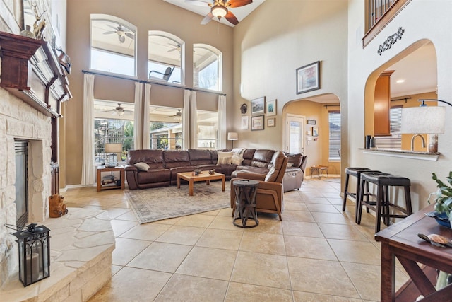 living room with light tile patterned floors, a ceiling fan, baseboards, arched walkways, and a stone fireplace