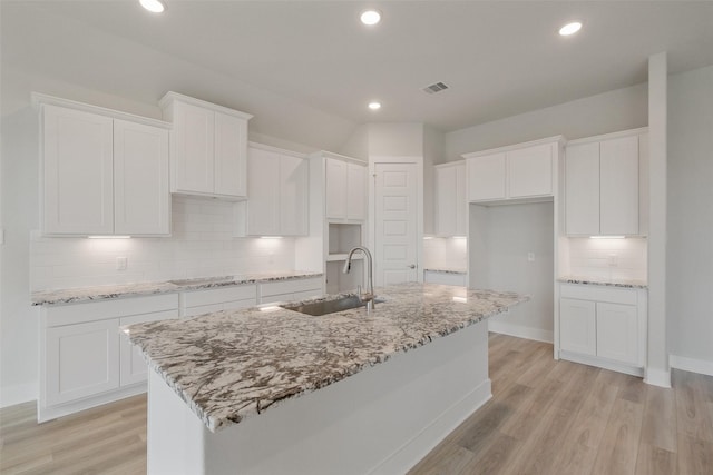 kitchen with light stone countertops, a center island with sink, a sink, white cabinetry, and light wood-type flooring