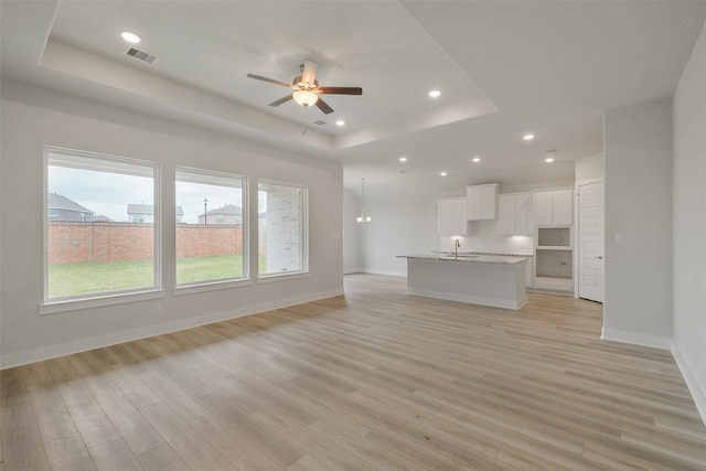 unfurnished living room with visible vents, ceiling fan with notable chandelier, a tray ceiling, light wood finished floors, and baseboards