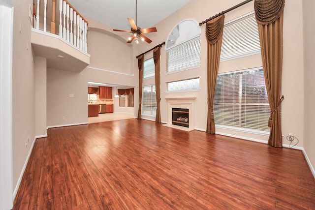 unfurnished living room featuring a fireplace with flush hearth, a ceiling fan, wood finished floors, baseboards, and a towering ceiling