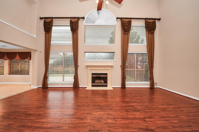 unfurnished living room featuring a fireplace with raised hearth, ceiling fan, baseboards, a high ceiling, and wood finished floors