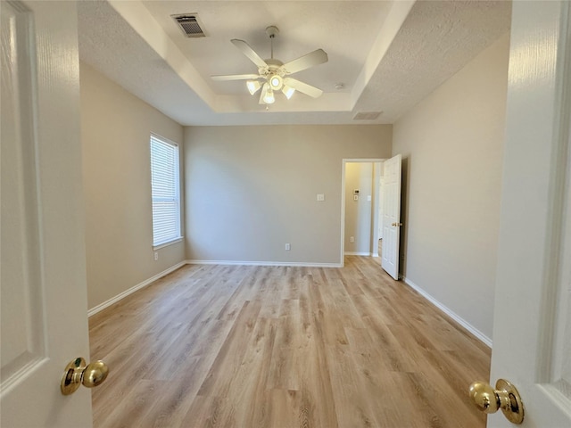 empty room with visible vents, light wood-style flooring, baseboards, and a tray ceiling