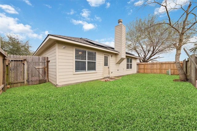 rear view of property with roof mounted solar panels, a chimney, a fenced backyard, a yard, and a gate