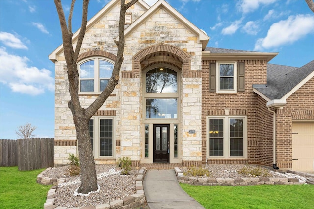 french country inspired facade featuring fence, roof with shingles, an attached garage, stone siding, and brick siding