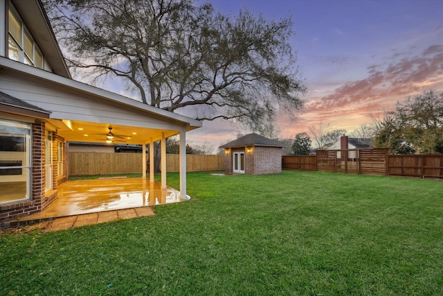 yard at dusk featuring a ceiling fan, an outbuilding, a fenced backyard, and a patio