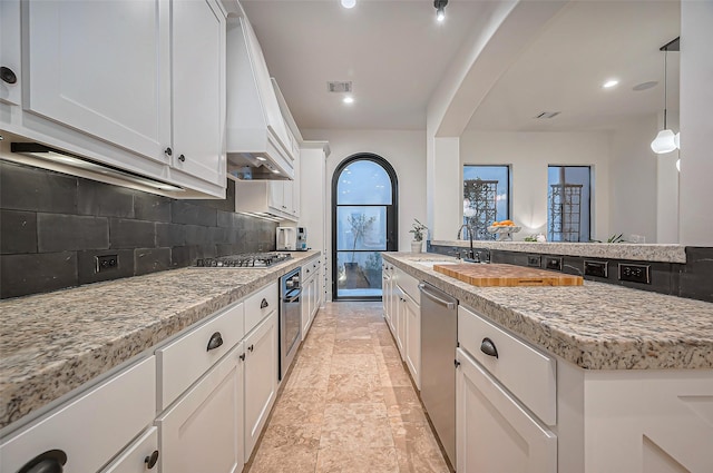 kitchen with custom range hood, decorative backsplash, stainless steel appliances, white cabinetry, and a sink