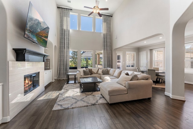 living area with baseboards, ceiling fan, a tiled fireplace, dark wood finished floors, and arched walkways