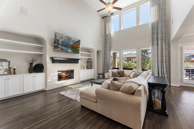 living area featuring built in shelves, baseboards, a tiled fireplace, a ceiling fan, and dark wood-style flooring