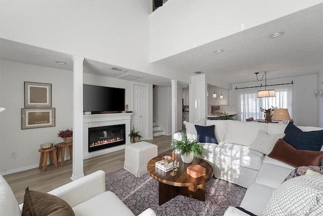 living room featuring visible vents, ornate columns, light wood finished floors, a high ceiling, and a glass covered fireplace