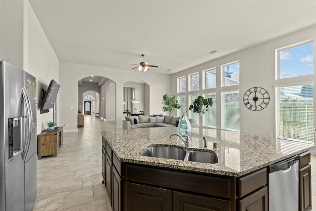 kitchen featuring dark brown cabinetry, a center island with sink, arched walkways, stainless steel appliances, and a sink