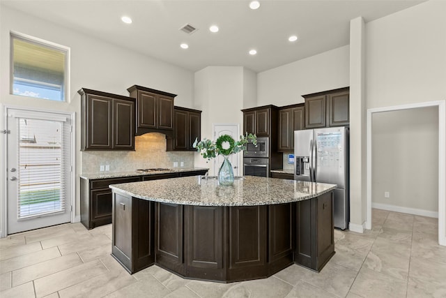 kitchen featuring light stone countertops, a high ceiling, a large island with sink, stainless steel appliances, and decorative backsplash