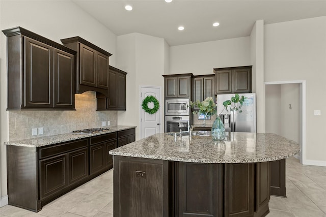 kitchen with light stone countertops, dark brown cabinetry, a center island with sink, decorative backsplash, and appliances with stainless steel finishes