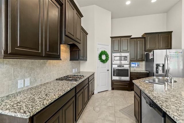 kitchen featuring tasteful backsplash, dark brown cabinets, light stone countertops, stainless steel appliances, and a sink