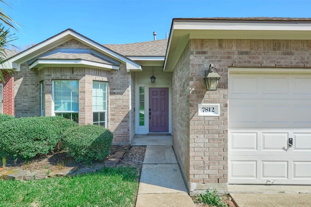 view of exterior entry with a garage, brick siding, and roof with shingles