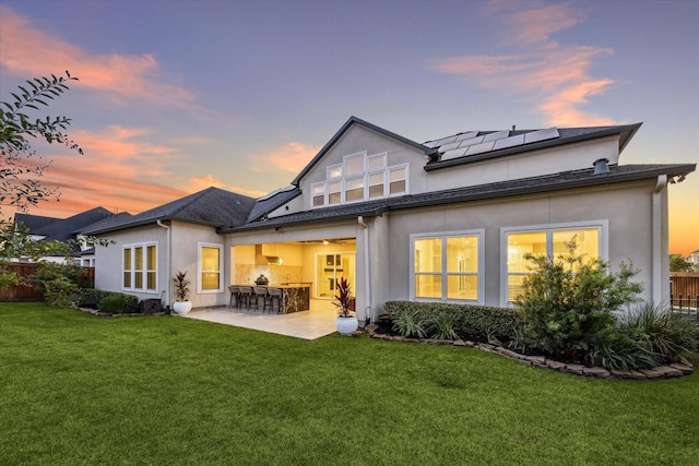 back of house at dusk with fence, solar panels, a patio area, a lawn, and outdoor dry bar