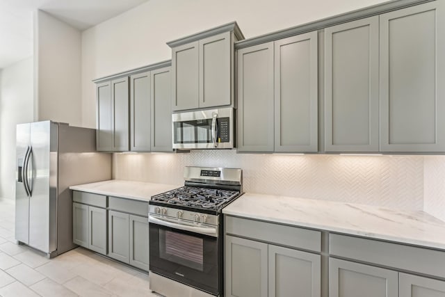 kitchen featuring gray cabinetry, tasteful backsplash, appliances with stainless steel finishes, light tile patterned flooring, and light stone countertops