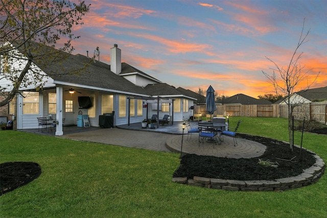 back of property at dusk with a lawn, a shingled roof, a patio, and fence