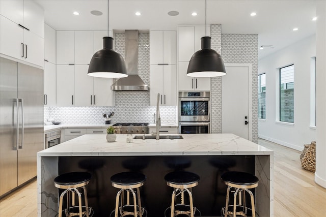 kitchen with light stone counters, stainless steel appliances, light wood-style floors, wall chimney range hood, and tasteful backsplash