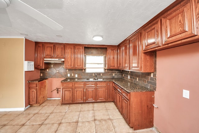 kitchen featuring under cabinet range hood, dark stone countertops, a sink, tasteful backsplash, and brown cabinetry