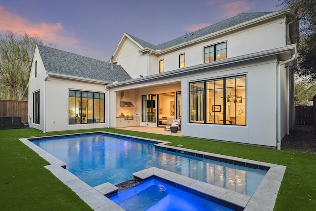 back of property at dusk featuring fence, exterior kitchen, a lawn, stucco siding, and an in ground hot tub