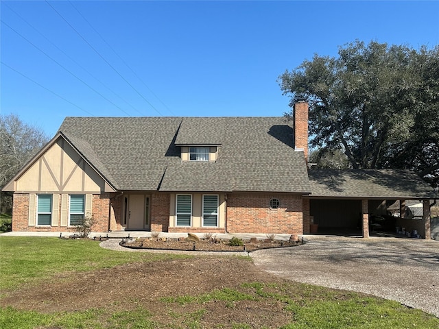 tudor home with gravel driveway, a front yard, a shingled roof, brick siding, and a chimney