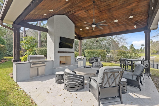 view of patio / terrace featuring ceiling fan, fence, exterior fireplace, an outdoor kitchen, and a grill