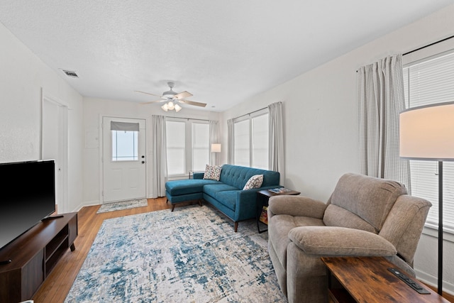 living room featuring a ceiling fan, baseboards, visible vents, light wood finished floors, and a textured ceiling