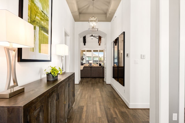 hallway with baseboards, an inviting chandelier, a tray ceiling, dark wood-style flooring, and arched walkways