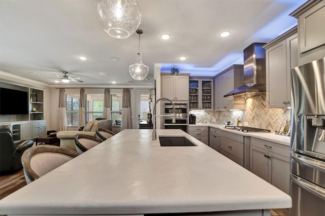 kitchen featuring a center island with sink, a sink, open floor plan, stainless steel appliances, and wall chimney range hood