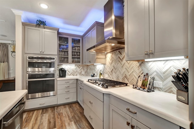 kitchen with wood finished floors, stainless steel appliances, light countertops, wall chimney range hood, and tasteful backsplash