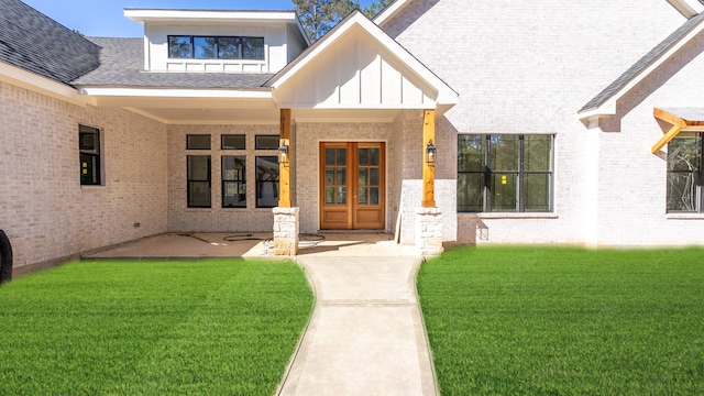 view of exterior entry with brick siding, a lawn, french doors, and a shingled roof