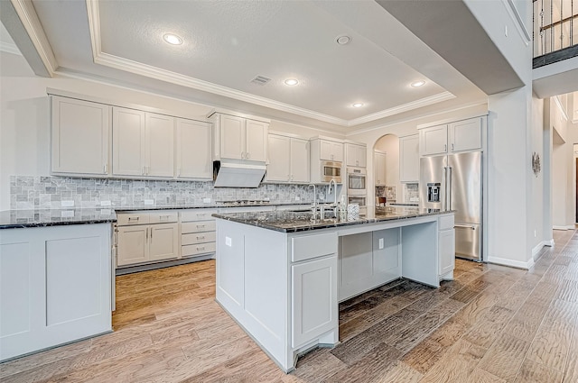 kitchen featuring a kitchen island with sink, stainless steel appliances, dark stone counters, crown molding, and light wood finished floors