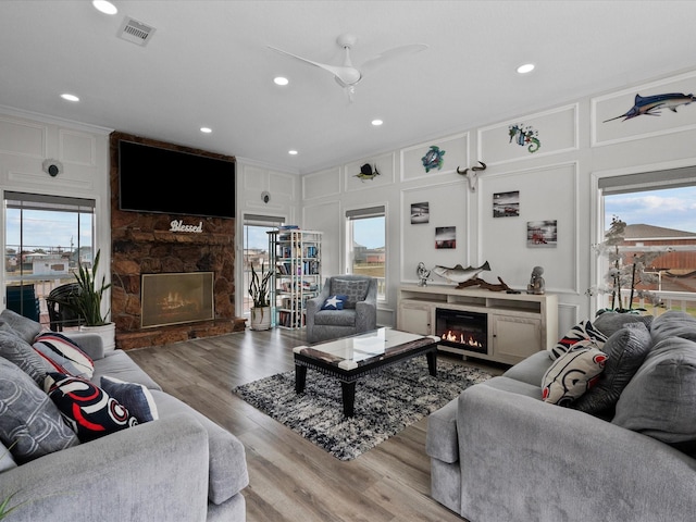 living room featuring a decorative wall, wood finished floors, visible vents, ceiling fan, and a stone fireplace