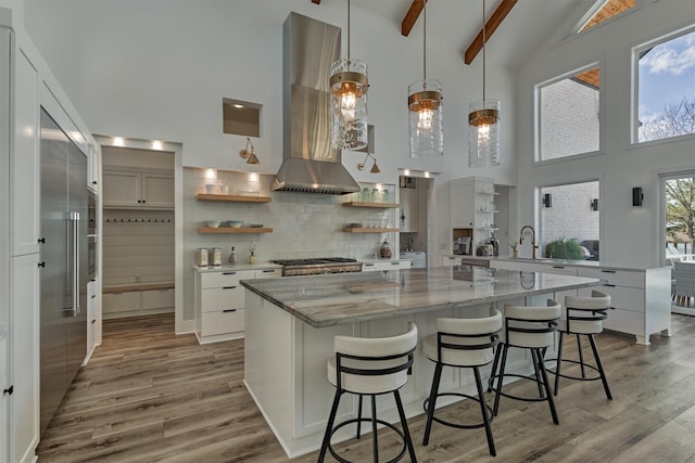 kitchen with open shelves, beamed ceiling, light wood-style flooring, and wall chimney range hood