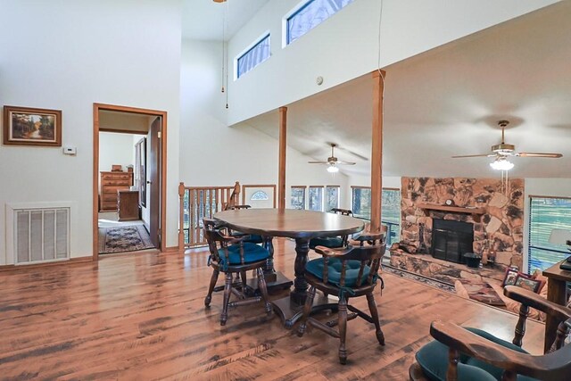 dining space featuring wood finished floors, visible vents, a ceiling fan, a stone fireplace, and a towering ceiling