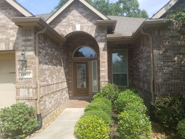 view of exterior entry with a garage, brick siding, and roof with shingles