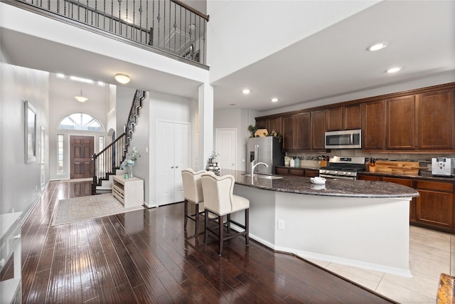 kitchen with dark stone counters, an island with sink, stainless steel appliances, a towering ceiling, and light wood-type flooring