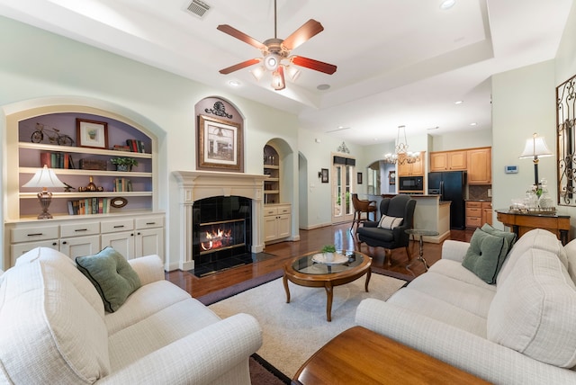 living room featuring a raised ceiling, wood finished floors, a fireplace with flush hearth, and ceiling fan with notable chandelier