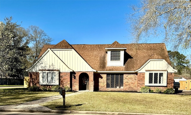 tudor-style house featuring a front lawn, fence, board and batten siding, roof with shingles, and brick siding