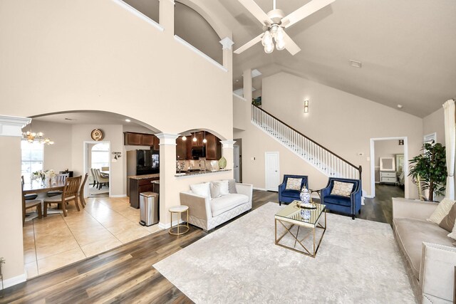 living area with light wood-type flooring, high vaulted ceiling, ceiling fan with notable chandelier, arched walkways, and decorative columns