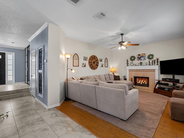 living room featuring light wood-type flooring, visible vents, a ceiling fan, a tiled fireplace, and a textured ceiling