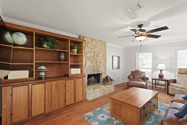 living area with visible vents, ceiling fan, a stone fireplace, light wood-style floors, and crown molding