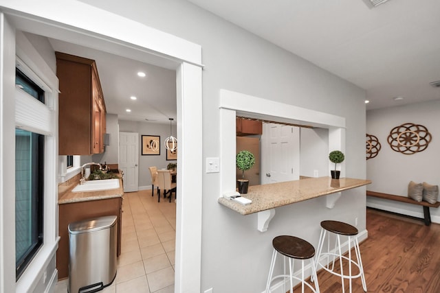 kitchen with a breakfast bar area, light stone counters, recessed lighting, brown cabinetry, and a sink