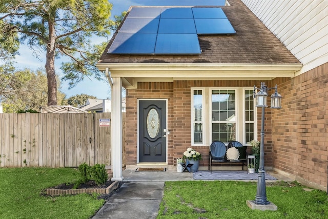 property entrance with brick siding, fence, roof with shingles, roof mounted solar panels, and a yard