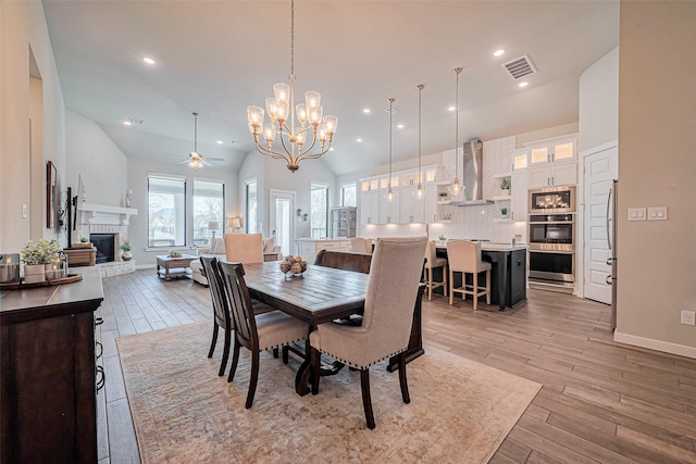 dining space featuring visible vents, high vaulted ceiling, light wood-style flooring, ceiling fan with notable chandelier, and a glass covered fireplace