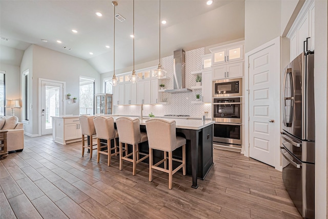 kitchen with an island with sink, wall chimney range hood, backsplash, stainless steel appliances, and light countertops